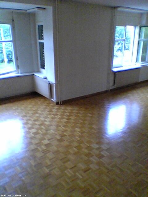 Pusta przestrzeń z parkietem ułożonym we wzór szachownicy, dwa okna z widokiem na zieleń na zewnątrz, ściany pomalowane na jasny kolor, widoczne rury grzewcze.